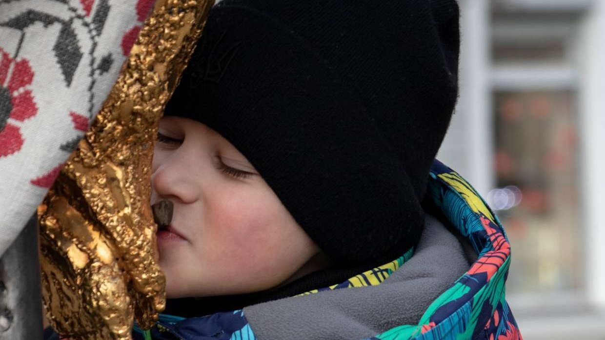 A child in winter clothes presses his face to the gilded foot of the Cross of Peace, Ukraine, winter 2025